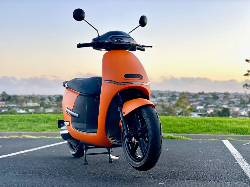 Orange electric scooter front view parked on a road with city background at sunset