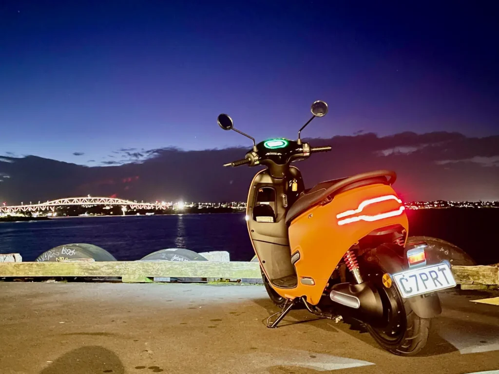 Orange electric scooter rear view parked near a waterfront at night with city lights
