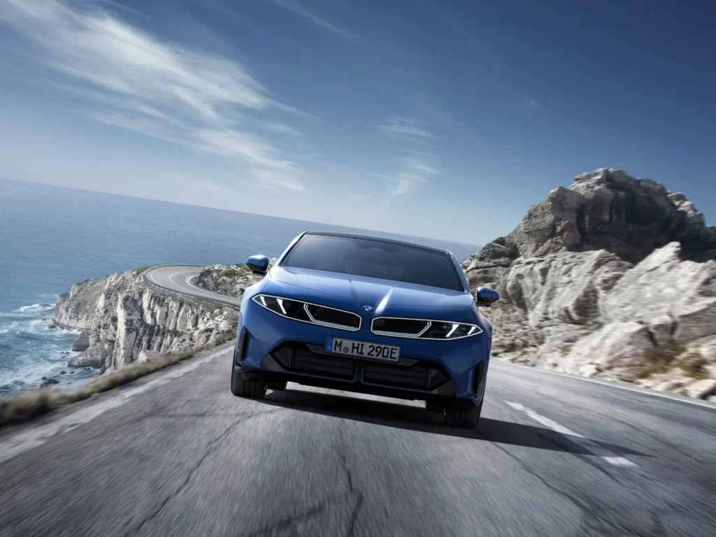 BMW i3 electric sedan front view driving toward camera on a coastal road with sea and cliffs in the background
