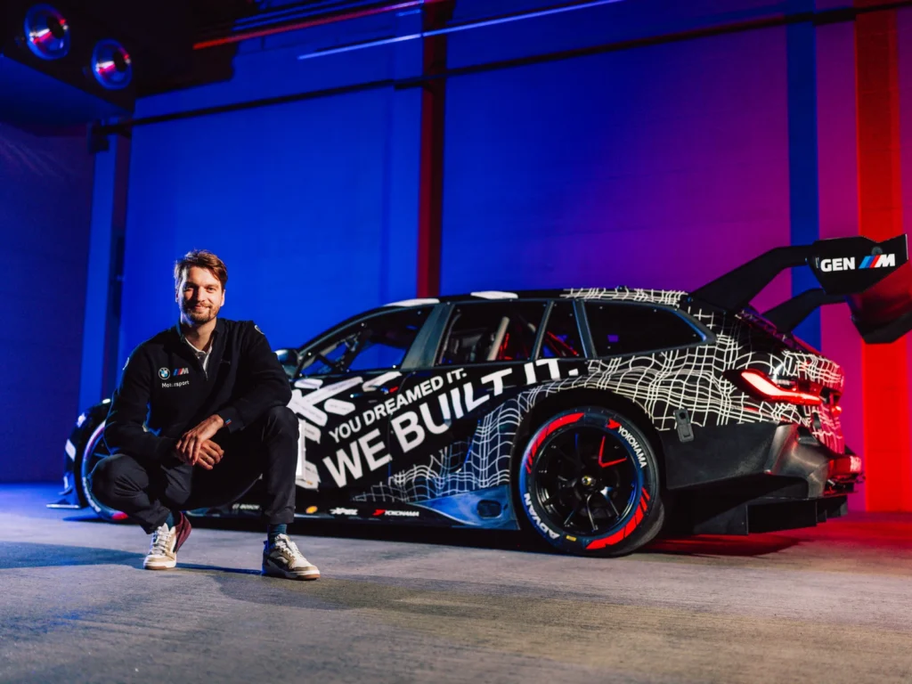 Man sitting beside BMW M3 Touring race car in studio with colorful lighting and branding